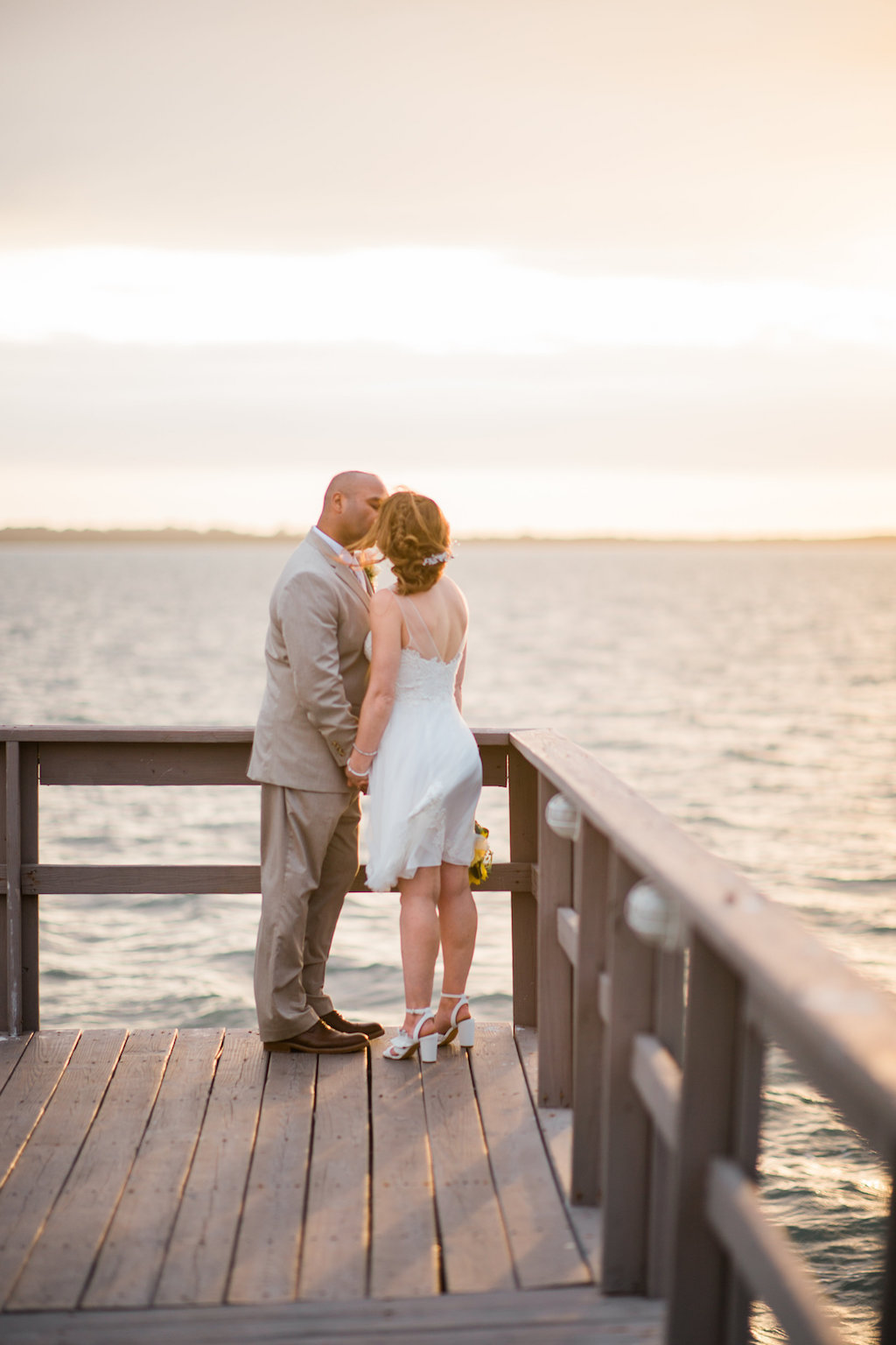 Intimate, Outdoor Sunset Waterfront Bride and Groom Wedding Portrait on Dock, Bride wearing White Tank Top Strap Short Reception Wedding Dress and Braid, Groom in Tan Suit and Blush Pink Rose Boutonniere | Dunedin Wedding Venue Beso Del Sol Resort