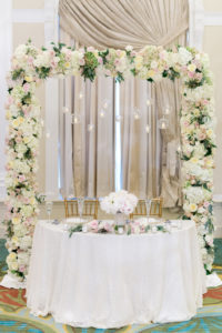 Wedding Reception Sweetheart Table Decor, Blush Pink, White and Greenery Floral Arch with Glass and Candle Spheres, Gold Chiavari Chairs, White Tablecloth, and Floral Bouquet Centerpiece