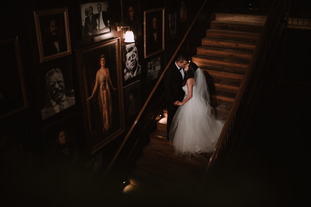 Indoor Wedding Portrait on Staircase, Bride in Sweetheart Strapless Tulle Wedding Dress with Rhinestone Belt and Cathedral Length Tulle Veil, Groom in Black Tuxedo with Black Bowtie and White Rose Boutonniere | Tampa Historic Wedding Venue The Oxford Exchange | Bridal Shop The Bride Tampa