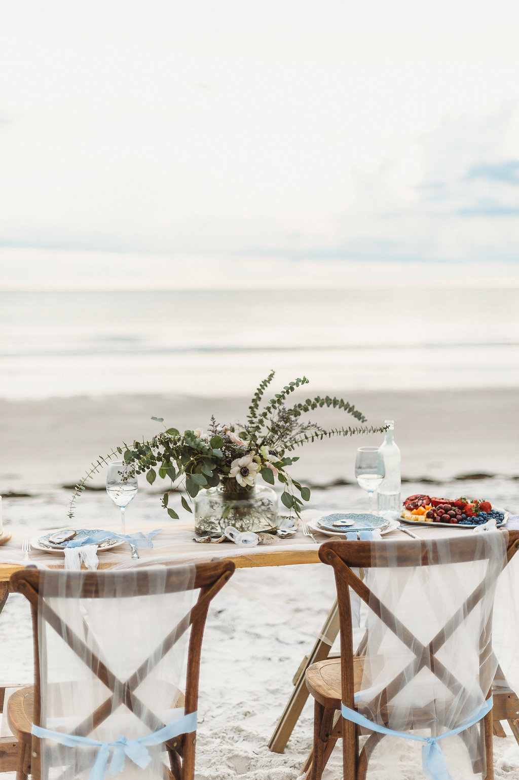 Waterfront, Coastal St. Pete Beach Wedding Reception Decor, Long Wooden Table, Wooden Crossback Chairs with Tulle and Light Blue Ribbon, Greenery and White Anemone Floral Centerpiece in Glass Vase