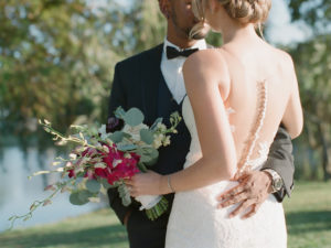 Intimate Outdoor Wedding Portrait, Bride in Spaghetti Strap Illusion Lowback Lace Dress with Buttons, Curled Hair Updo and Pink, Deep Purple, White and Greenery Floral Bouquet, Groom in Black Tuxedo and Black Bowtie