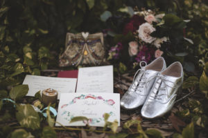 Old English Floral Inspired Wedding, White and Floral Invitation, Wedding Rings on Wooden Log, Men Silver Wingtip Shoes, Blush Pink, Dark Maroon and Greenery Bouquet on Leaves