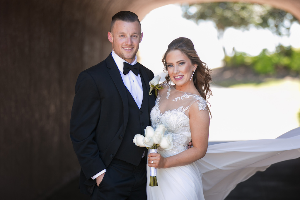Outdoor Bride and Groom Wedding Portrait Underneath Greenery Bridge, Bride in Maggie Sottero Illusion Tank Top with Beaded Lace Motif Wedding Dress with White Rose Bouquet, Groom in Black Tuxedo | Sarasota Wedding Photographer Carrie Wildes Photography