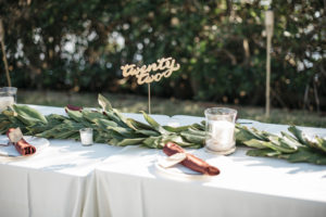 Outdoor Wedding Reception Decor Centerpieces Long Feasting Table with White Tablecloth, Long Greenery Garland, Burgundy Linens, Clear Class Hurricane Vase with Candle and Gold Lasercut Table Number | Tampa Bay Wedding Planner Kelly Kennedy Weddings and Events