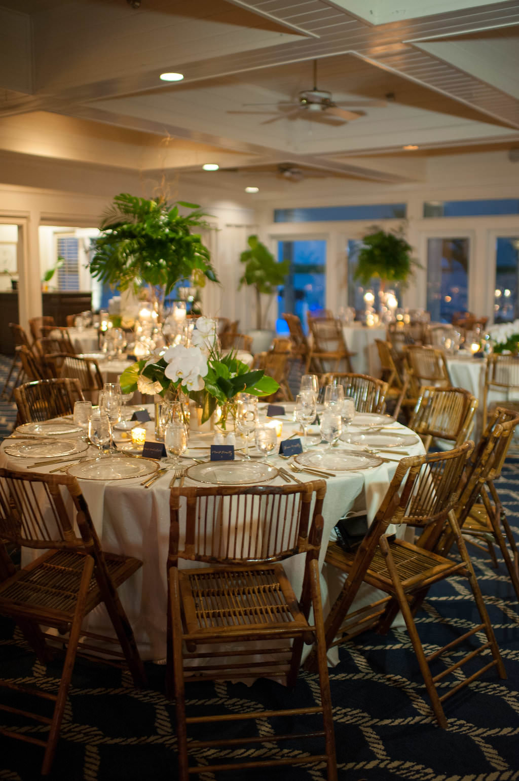 Indoor Wedding Reception Decor, Tall Glass Cylinder Vase with Tropical Green Leaves and Low Centerpiece with White Orchid Florals and Greenery and Wooden Brown Chairs | Tampa Wedding Planner Parties A La Carte | Clearwater Beach Wedding Venue Carlouel Yacht Club | St Pete Rentals Over The Top Linen Rentals and A Chair Affair