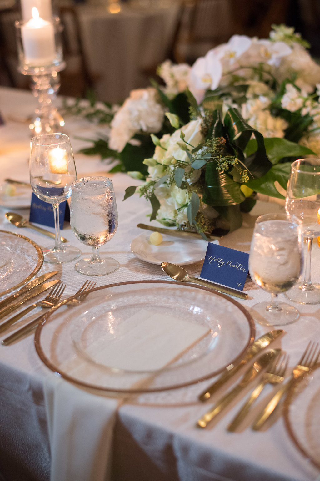 Indoor Wedding Reception Sweetheart Table Decor, Low Centerpiece with White Orchid Floral and Tropical Greenery Leaves and Clear Glass and Gold Rim Charger on White Linen | Tampa Wedding Planner Parties A La Carte | St Pete Rentals Over The Top Linen Rentals