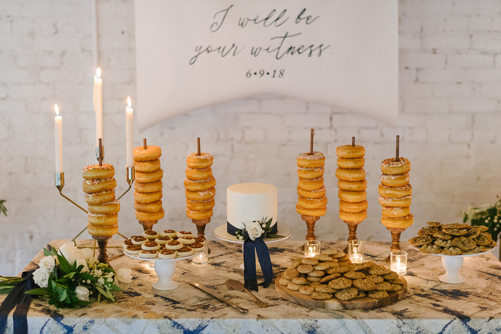 Rustic Wedding Reception Dessert Table with Cookies and Mini Doughnuts, Candlestick, One Tier White Wedding Cake with Navy Blue Ribbon on Artsy Blue and White Linen, White and Greenery Bouquet and White Fabric Wedding Banner | Lakeland Wedding Venue Haus 820 | Tampa Bay Wedding Rentals Over the Top Linens