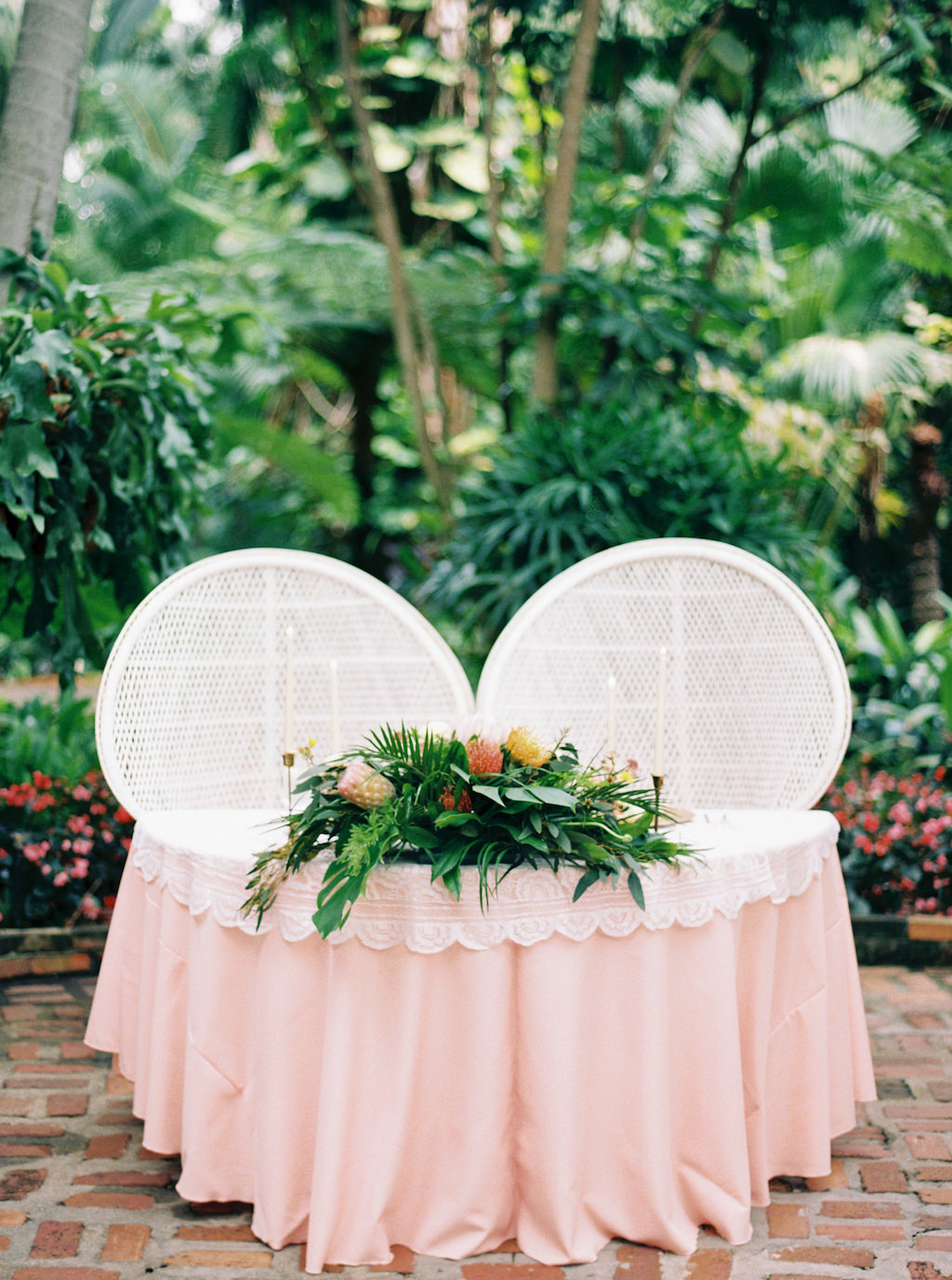Outdoor Tropical Inspired Wedding Reception Decor, Sweetheart Table with Blush Pink and White Lace Tablecloth, Large White Wicker Chairs, and Palm Leaves, Pink and Yellow Floral Bouquet