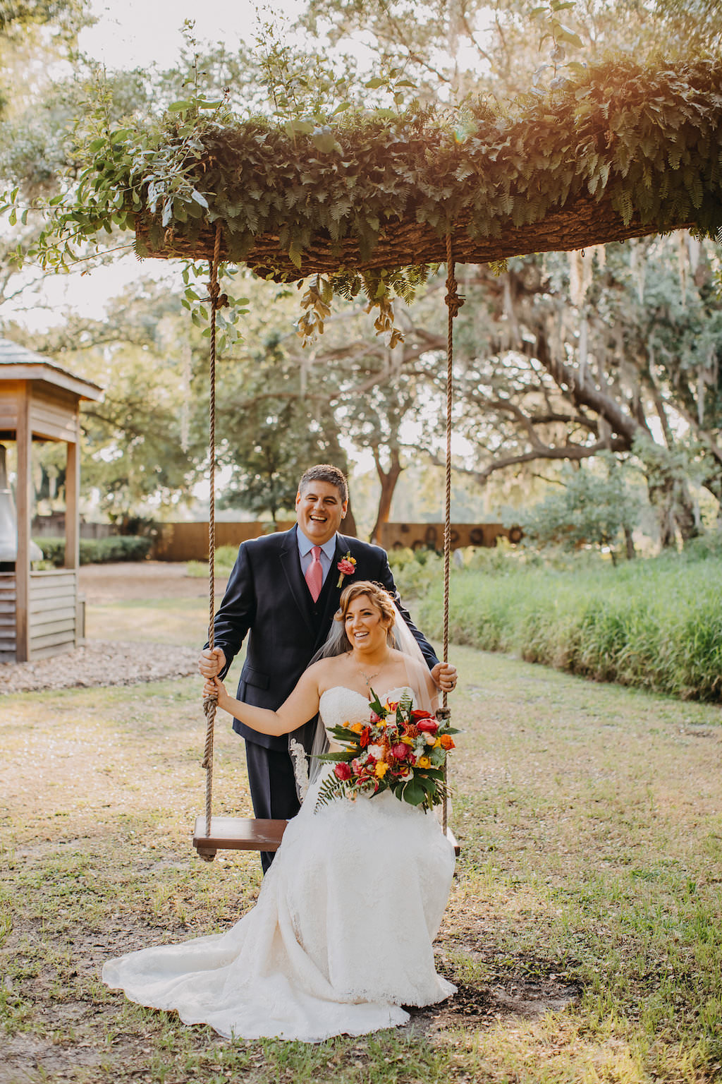 Outdoor Bride and Groom Wedding Portrait on Swing from Tree, Bride in A-Line Sweetheart Strapless Lace Wedding Dress with Tropical Floral Bouquet, Groom in Navy Blue Suit and Pink Tie | Tampa Bay Photographer Rad Red Creative | Rustic Venue Cross Creek Ranch