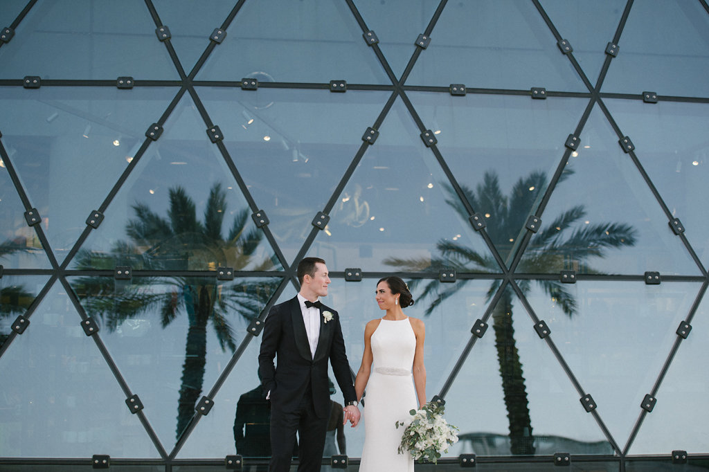 Outdoor Bride and Groom Wedding Portrait in Front of Unique Geometric Windows, Bride in Sleek High Neckline Wedding Dress with Rhinestone Belt and White and Greenery Bouquet, Groom in Black Tuxedo | Tampa Bay Hair and Makeup Femme Akoi