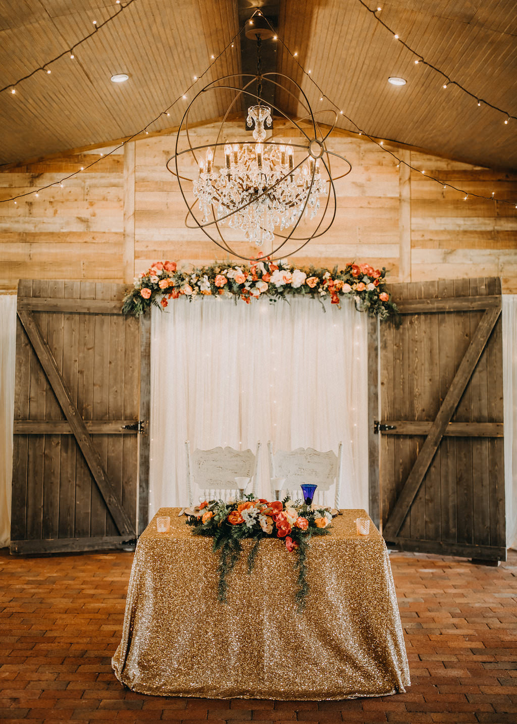 Glamorous Rustic Wedding Reception Decor, Sweetheart Table with Gold Sparkle Tablecloth, Tropical Floral Bouquet, White Wooden Chairs, Linen Drape Backdrop with Red, Orange, Pink, White and Greenery Flowers, String Lights and Chandelier | Tampa Bay Photographer Rad Red Creative | Rustic Venue Cross Creek Ranch