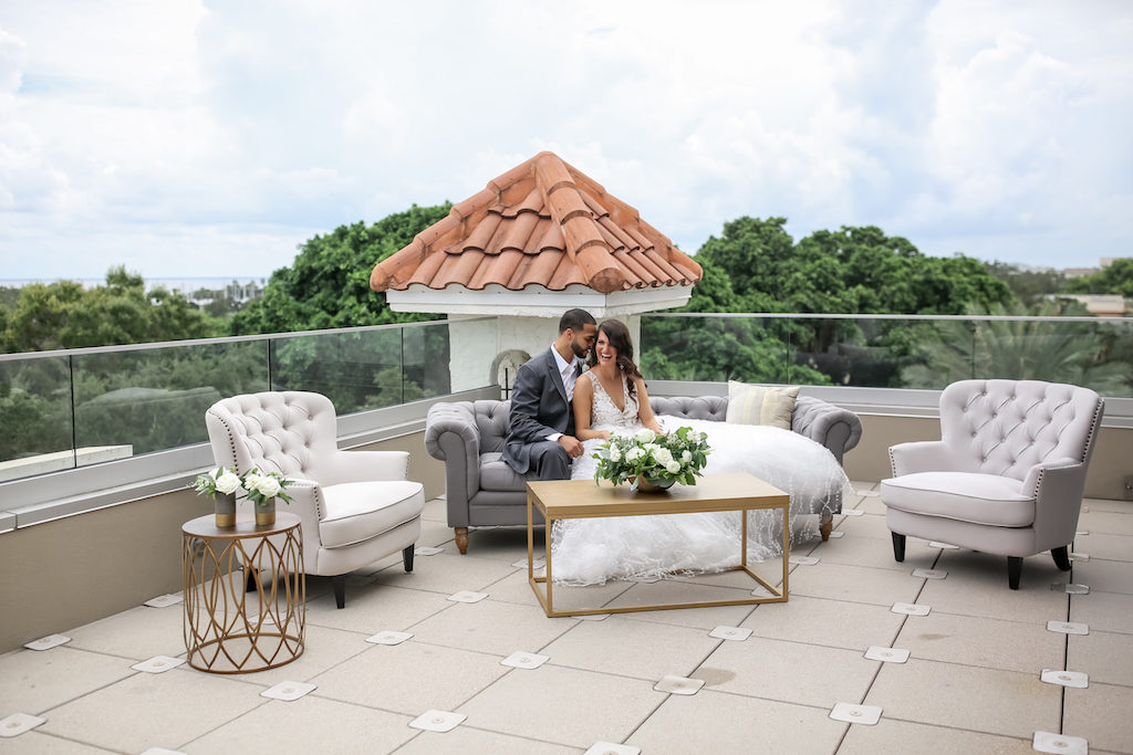 Bride and Groom Hotel Rooftop Wedding Portrait on Grey Modern Tufted Couch, Gold Coffee Table with White and Greenery Floral Centerpiece, White Tufted Armchairs, Gold Side Table with White Florals in Vases | Tampa Bay Photographer Lifelong Photography Studios | Planner Parties A'la Carte | Florist Cotton and Magnolia | Downtown St. Pete Wedding Venue The Birchwood | Rentals A Chair Affair