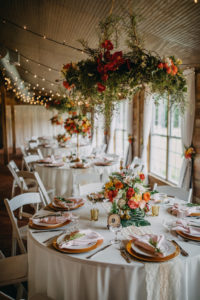 Tropical Inspired Wedding Reception Decor, Round Tables with White Folding Chairs, White Tablecloth, Gold Chargers, Blush Pink Linens, Low Tropical Pink, Yellow, Orange, White and Greenery Floral Bouquet, String Lights and Orange, Red, and Greenery Floral Bouquet Hanging from Ceiling | Tampa Bay Photographer Rad Red Creative | Rustic Venue Cross Creek Ranch