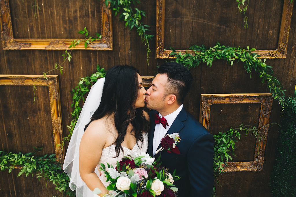 Outdoor Bride and Groom First Look Wedding Portrait, Bride in Sweetheart Strapless Rhinestone Beaded Wedding Dress with Veil, Greenery, Ivory, Pink and Red Bouquet, Groom in Navy Blue Tuxedo with Red Bowtie, Red Pocket Square, and Plum Boutonniere