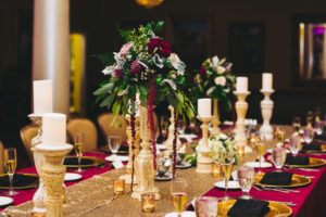 Vintage Modern Wedding Reception Decor, Long Feasting Table with Red Tablecloth, Gold Chargers, Black Linens, Tall Wooden Candlesticks, Dark Red, Pink, White and Greenery Floral Bouquet with Marsala Red Hanging Amaranthus on Tall Wooden Vintage Holder on Gold Table Runner