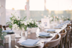 Long Wooden Feasting Table with Wood Crossback Chairs, Low White Vases with White and Greenery Floral Centerpieces, Silver Candle Votives, and Pale Blue Napkin Linen | Tampa Bay Rental Company A Chair Affair