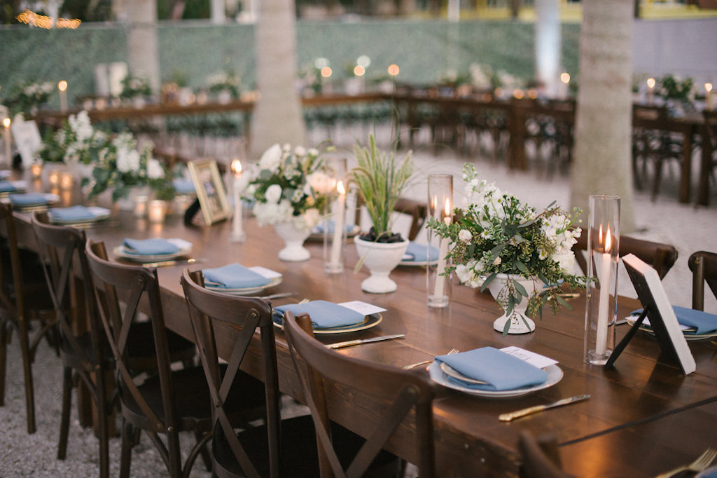 Long Wooden Feasting Table with Wood Crossback Chairs, Low White Vases with White and Greenery Floral Centerpieces, Tall Glass Vases with Candlesticks, Picture Frames, and Pale Blue Napkin Linens on Antique China Plates | Tampa Bay Rental Company A Chair Affair | Unique St. Petersburg Venue Intermezzo Coffee and Cocktails