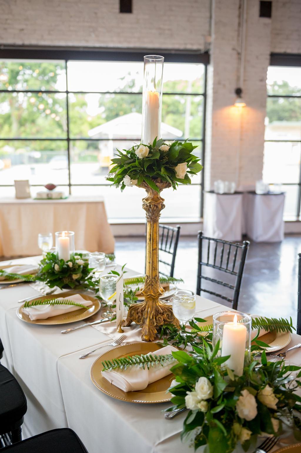 Wedding Reception Decor, Long Table with White Tablecloth, Gold Silk Table Runner, Gold Chargers, White Linen, Green Leaf, Tall Gold Candlestick with White Rose and Greenery Topper and Glass Cylinder Candle Holder and Black Chiavari Chairs | Tampa Bay Photographer Andi Diamond Photography | Rentals A Chair Affair