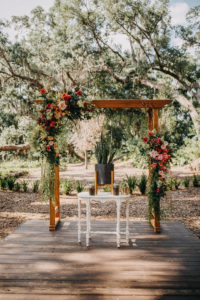 Outdoor Tropical Inspired Wedding Ceremony Decor, Wooden Arch with Pink, Yellow, Orange, Red and Greenery Bouquets, White Ceremony Table with Black and Gold Vase withe Palm Tree Leaves | Tampa Bay Photographer Rad Red Creative | Venue Cross Creek Ranch
