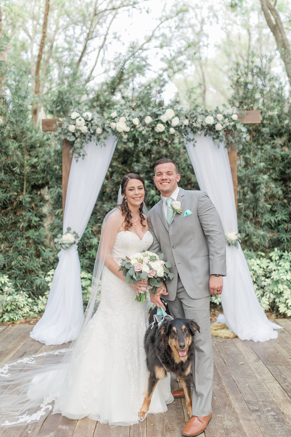 Outdoor Rustic Elegant Bride, Groom and Dog Wedding Portrait, Bride in Sweetheart Strapless Lace Fit and Flare Wedding Dress, Groom in Grey Suit, Wooden Arch with White Linen Draping with Ivory Roses and Greenery Garland | Plant City Wedding Venue Florida Rustic Barn Weddings