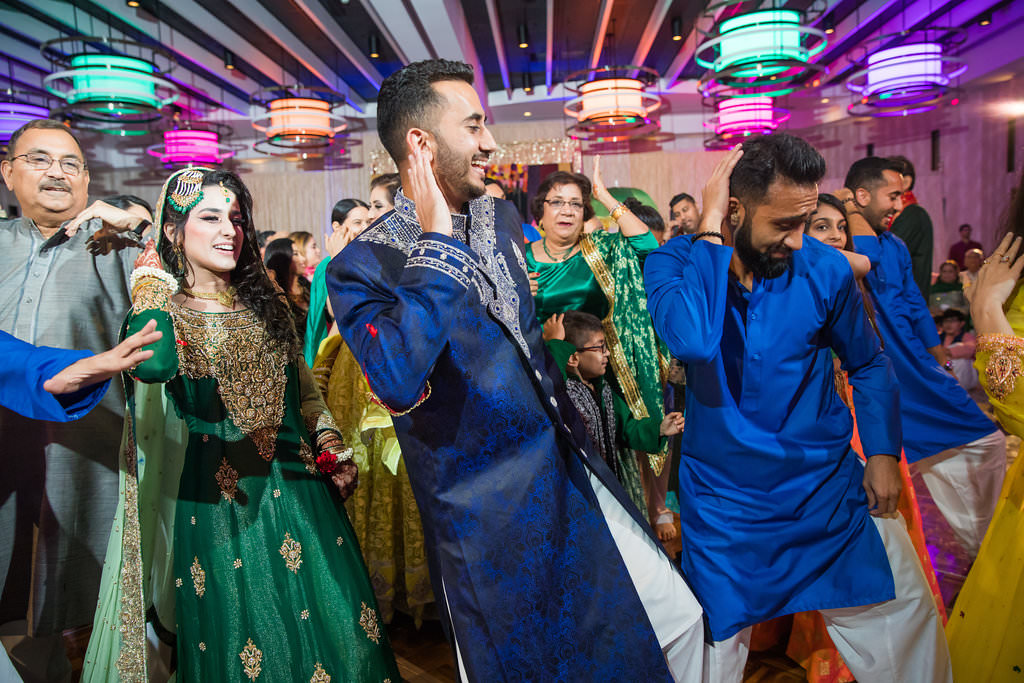 Glamorous Traditional Indian Wedding Reception Dancing, Bride in Gold and Green Sari, Groom in Blue and White Traditional Indian Attire with Colorful Chandelier Lighting