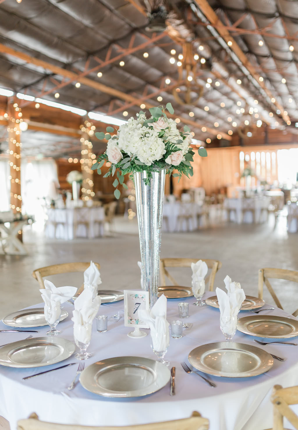 Rustic Elegant Tampa Bay Barn Reception Decor, Round Tables with Grey Tablecloths, Silver Chargers, Tall Blush Pink Roses, White Hydrangeas and Greenery Floral Centerpiece in Silver Vase, Table Number in White Frame, Wood Chiavari Chairs | Plant City Wedding Venue Florida Rustic Barn Weddings