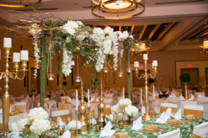 Glamorous Traditional Indian Wedding Hotel Ballroom Reception Decor, Long Feasting Table, Green Decorative Tablecloth, Chairs with White Covers and Gold Sashes, Tall Gold Candlesticks Centerpieces, White Floral, Hanging Amaranthus and Wooden Branches with Hanging Glass Cylinders with Candles | Tampa Bay Wedding Venue Wyndham Grand Clearwater Beach