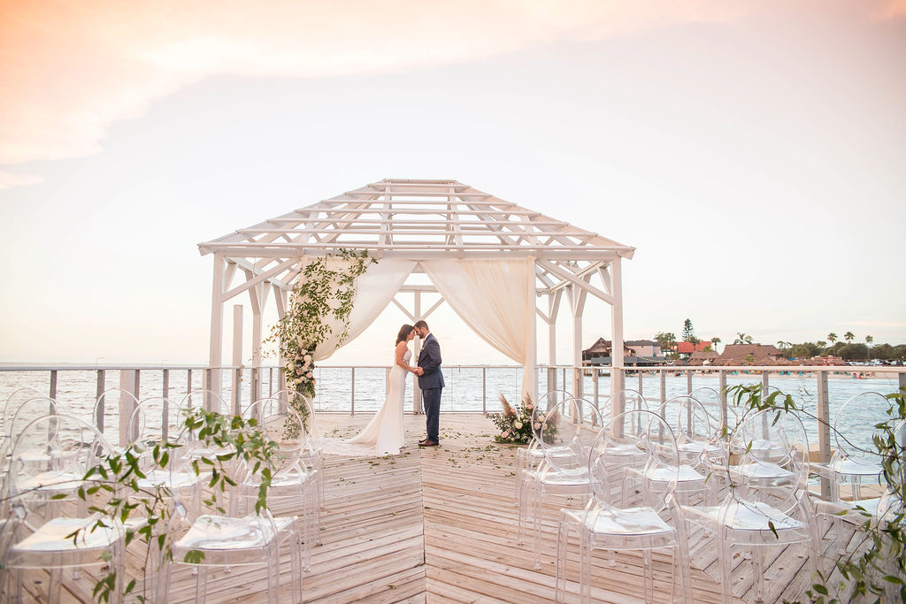 Florida Outdoor Waterfront Wedding Ceremony Bride and Groom Portrait Under Wooden White Canopy with Arch Draped with White Linen, Greenery Leaves and Ivory and Blush Pink Florals, Acrylic Chairs with Greenery Garland | Tampa Wedding Photographer Kristen Marie Photography | Tampa Venue The Godfrey | Draping and Ghost Chair Rentals Gabro Event Services Instagram