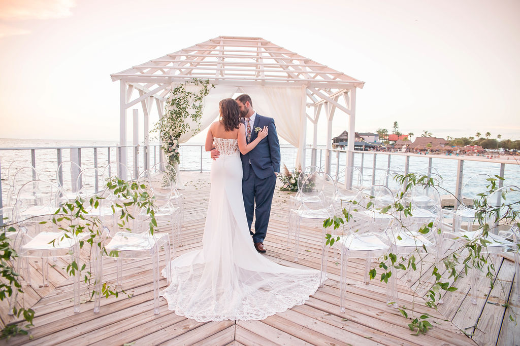 Florida Outdoor Waterfront Wedding Ceremony Bride and Groom Portrait Under Wooden White Canopy with Arch Draped with White Linen, Greenery Leaves and Ivory and Blush Pink Florals, Acrylic Chairs with Greenery Garland | Tampa Wedding Photographer Kristen Marie Photography | Tampa Venue The Godfrey | Draping and Ghost Chair Rentals Gabro Event Services