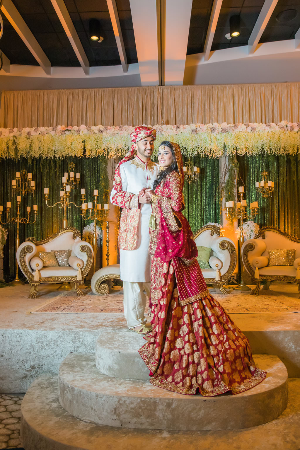 Glamorous Traditional Indian Bride and Groom Wedding Portrait, Bride in Gold and Red Sari, Groom in White Indian Wedding Attire and Red and Gold Turban, Green Linen Backdrop with Hanging White Florals, Ivory and Gold Couch Lounge Seating and Gold Candlesticks | Tampa Bay Wedding Venue Wyndham Grand Clearwater Beach