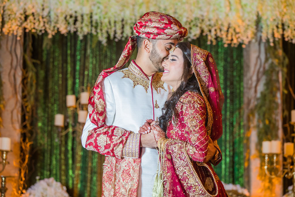 Glamorous Traditional Indian Bride and Groom Wedding Portrait, Bride in Gold and Red Sari, Groom in White Indian Wedding Attire and Red and Gold Turban, Green Linen Backdrop with Hanging White Florals, Ivory and Gold Couch Lounge Seating and Gold Candlesticks | Tampa Bay Wedding Venue Wyndham Grand Clearwater Beach