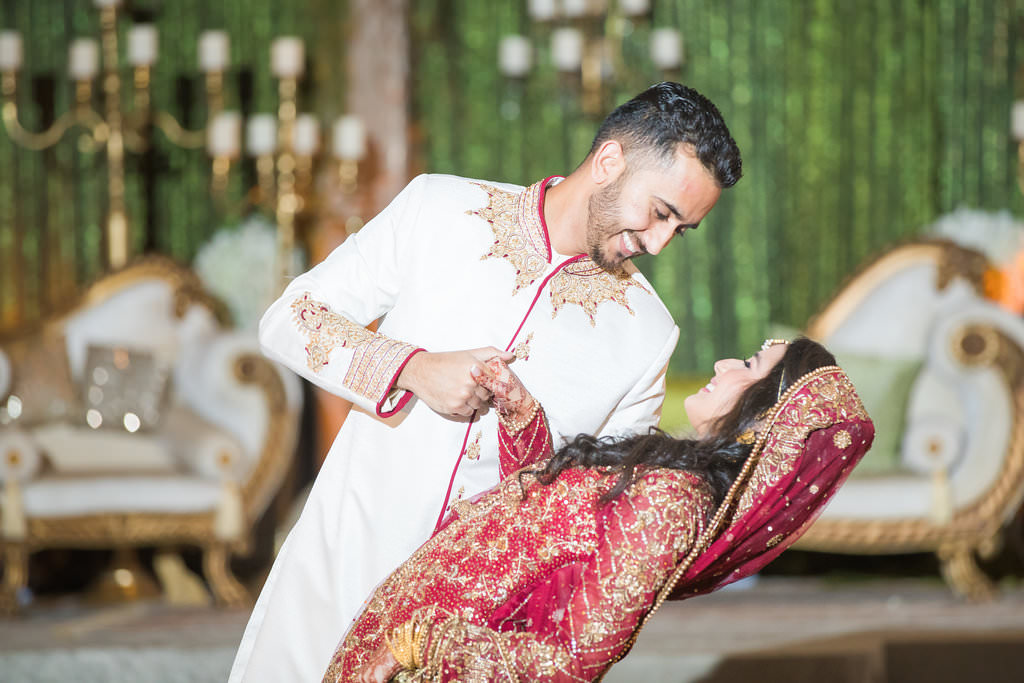 Glamorous Traditional Indian Bride and Groom Wedding Portrait, Bride in Gold and Red Sari, Groom in White Indian Wedding Attire and Red and Gold Turban, Green Linen Backdrop with Hanging White Florals, Ivory and Gold Couch Lounge Seating and Gold Candlesticks | Tampa Bay Wedding Venue Wyndham Grand Clearwater Beach