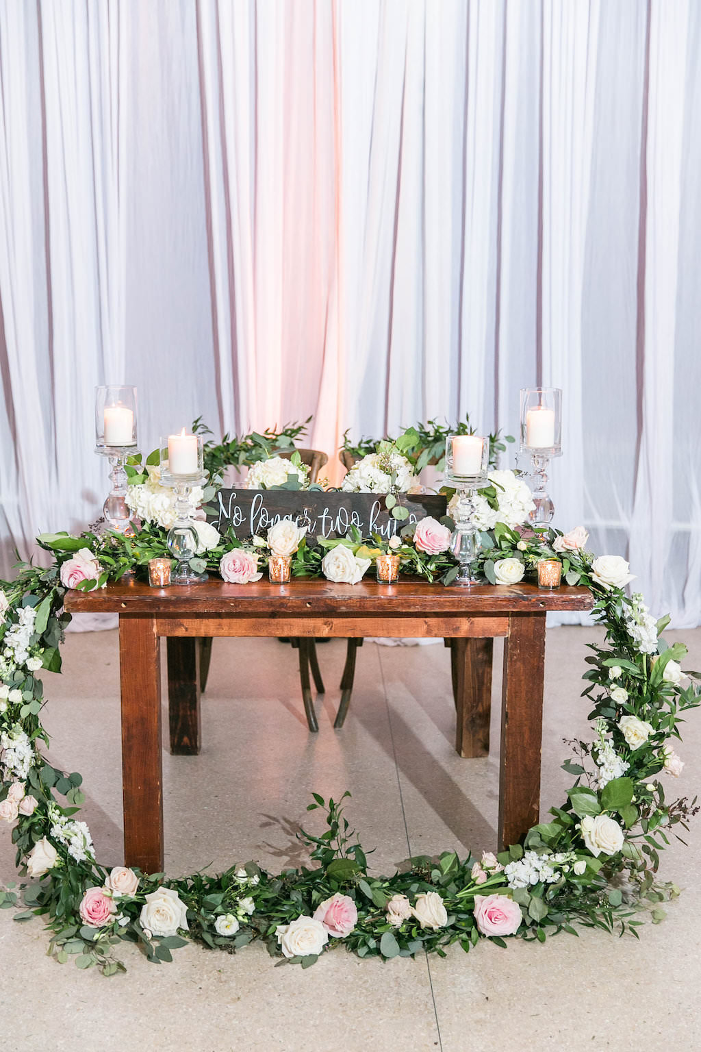 Wedding Reception Garden Inspired Decor, Wooden Sweetheart Table with Circular Greenery Garland with Ivory and Blush Pink Roses, Tall Glass Candlesticks and Wooden Sign | Tampa Bay Rentals A Chair Affair