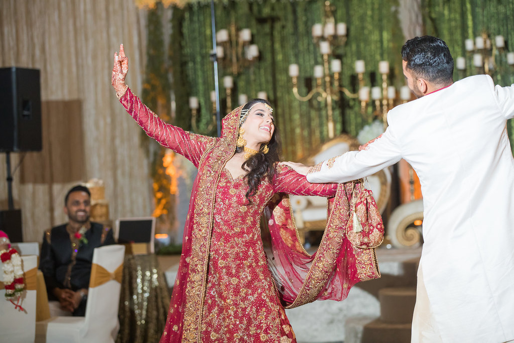 Glamorous Traditional Indian Bride and Groom Wedding Portrait, Bride in Gold and Red Sari, Groom in White Indian Wedding Attire and Red and Gold Turban, Green Linen Backdrop with Hanging White Florals, Ivory and Gold Couch Lounge Seating and Gold Candlesticks | Tampa Bay Wedding Venue Wyndham Grand Clearwater Beach