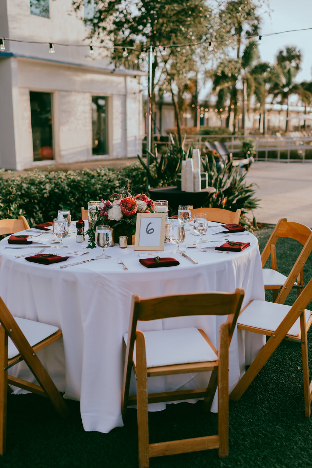 Outdoor Garden Lawn Wedding Reception Decor, Round Table, White Tablecloths, Burgundy Linens, Wooden Folding Chairs with White Cushions, Gold Frame Table Number and Low Ivory, Red and Greenery Floral Bouquet Centerpiece | St. Petersburg Wedding Venue Postcard Inn on the Beach