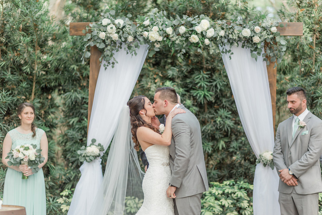 Outdoor Rustic Elegant Tampa Bay Wedding Ceremony Portrait, Bride and Groom First Kiss, Wooden Arch with White Linen Draping, Ivory Roses and Greenery Garland | Plant City Wedding Venue Florida Rustic Barn Weddings