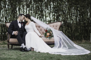 Outdoor Florida Bride and Groom Wedding Portrait on Blush Pink Vintage Loveseat Couch, Bride in Crepe and Lace Long Sleeve V Neckline Mermaid Wedding Dress with Cathedral Veil, Groom in Black Tuxedo