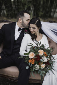 Outdoor Florida Bride and Groom Wedding Portrait on Blush Pink Vintage Loveseat Couch, Bride in Crepe and Lace Long Sleeve V Neckline Mermaid Wedding Dress with Cathedral Veil with Greenery, Blush Pink, Orange, and Red Floral Bouquet, Groom in Black Tuxedo