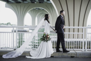 Outdoor St. Petersburg Florida Bride and Groom Wedding Portrait Under Intracoastal Waterway Bridge, Bride in Mermaid Crepe and Lace Long Sleeve Wedding Dress and Cathedral Veil with Greenery, Red, Blush Pink Floral Bouquet, Groom in Black Tuxedo