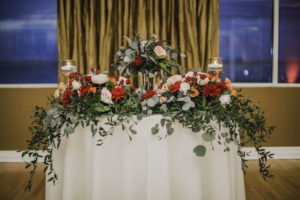 Romantic, Florida Ballroom Reception Wedding Decor, Sweetheart Table with White Linen, Greenery, Red, Orange and Blush Floral Garland and Floral Bouquet in Tall Glass Vase Centerpiece