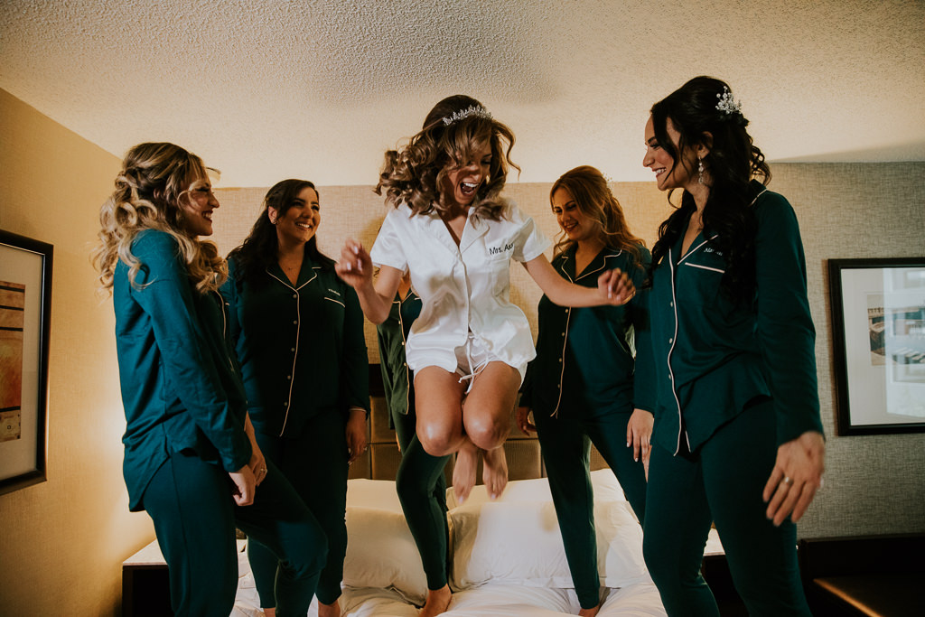 Bride and Bridesmaids Getting Ready Wedding Portrait Jumping on Bed, Bridesmaids in Matching Green Long Sleeve Pajamas, Bride in Short Sleeve and Shorts White Pajamas | Tampa Bay Wedding Photographer Brandi Image Photography | Downtown Tampa Hotel Wedding Venue Tampa Hilton Downtown