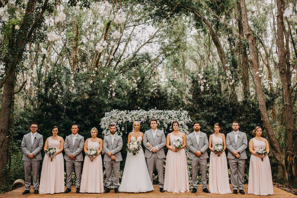 Florida Outdoor Rustic Inspired Wedding, Bride, Groom and Wedding Party Portrait, Bridesmaids in Matching One Shoulder Blush Pink Dresses, Groomsmen in Grey Suits, Bride in Spaghetti Strap V Neck Lace A Line Wedding Dress, Greenery and White Floral Arch with Hanging Glass Bulbs | Lakeland Rustic Wedding Venue The Prairie Glenn Barn at Gable Oaks Ranch