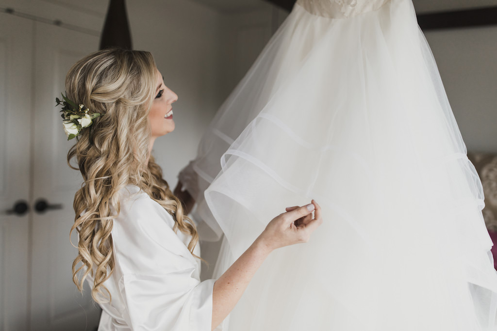 Florida Bride Getting Ready Wedding Portrait, Bride in White Silk Robe with Boho Chic Hair, Curls and Real White and Greenery Floral Headpiece Looking at Wedding Dress | Tampa Bay Wedding Hair and Makeup Artist Michele Renee the Studio
