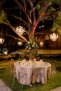 Nighttime Outdoor Garden Courtyard Wedding Reception Decor, Round Tables with Ivory Tablecloths, Ghost Chairs, Tall Greenery, White, Ivory, Red Floral Centerpiece, Crystal Chandeliers Hanging From Tree | Sarasota Wedding Venue Ringling Museum | Tampa Bay Wedding Planner NK Weddings