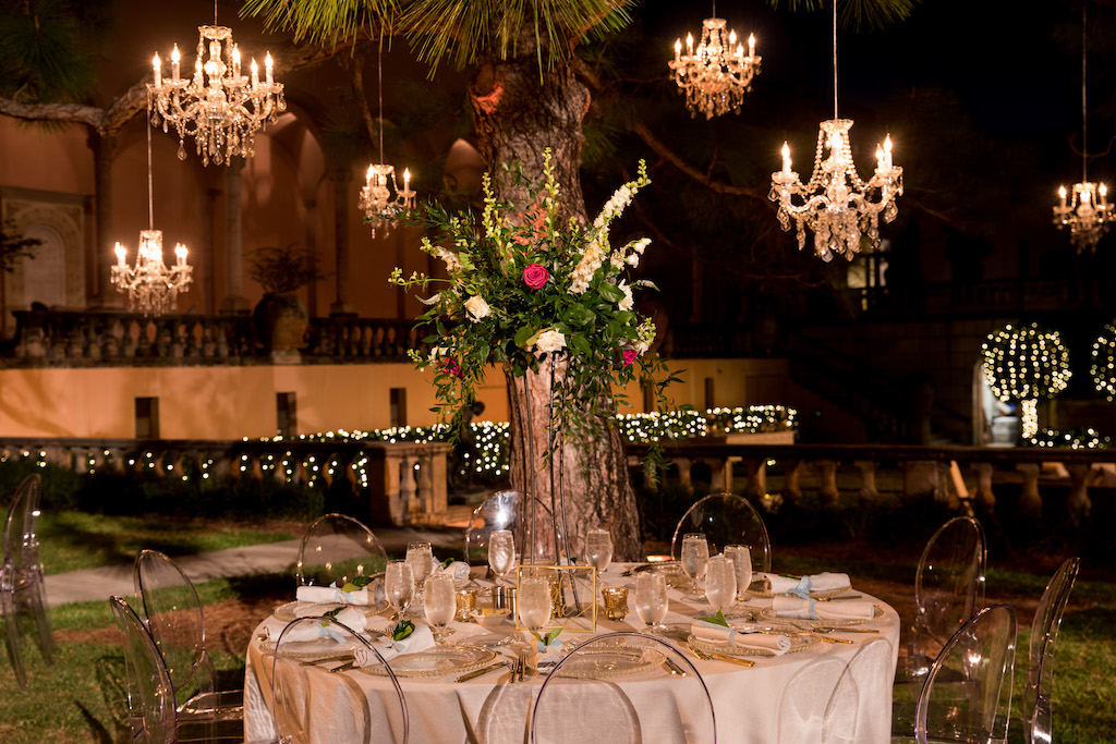 Nighttime Outdoor Garden Courtyard Wedding Reception Decor, Round Tables with Ivory Tablecloths, Ghost Chairs, Tall Greenery, White, Ivory, Red Floral Centerpiece, Crystal Chandeliers Hanging From Tree | Sarasota Wedding Venue Ringling Museum | Tampa Bay Wedding Planner NK Weddings