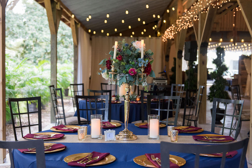 Rustic Wedding Reception Decor, Round Tables with Navy Blue Tablecloths, Gold Chargers and Burgundy Linens, Tall Silver Candlestick with Greenery, Ivory and Red Floral Bouquet Centerpiece, Gold Table Runner, Bistro Hanging Lights | Rustic Tampa Wedding Venue Kathleen's Garden