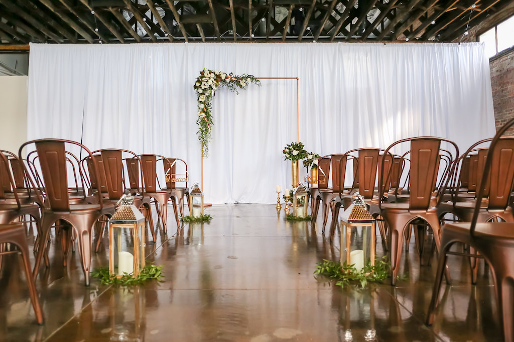 Industrial Inspired Wedding Ceremony, Bronze Metal Arch with White and Greenery Florals, Bronze Metal Chairs, Gold Lanterns with Greenery, White Draping | Tampa Bay Wedding Photographer Lifelong Photography Studios | Tampa Unique Industrial Wedding Venue CAVU | Wedding Rentals A Chair Affair