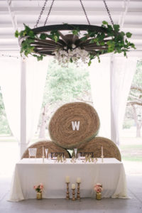 Tampa Bay Rustic Wedding Reception Decor, Sweetheart Table with White Tablecloth, Gold Candlesticks, Blush Pink Floral Arrangements in Gold Vases, Gold Mr and Mrs Signs, Hay Barrels Backdrop, Wooden Wheel Chandelier Decorated with Greenery and White Florals
