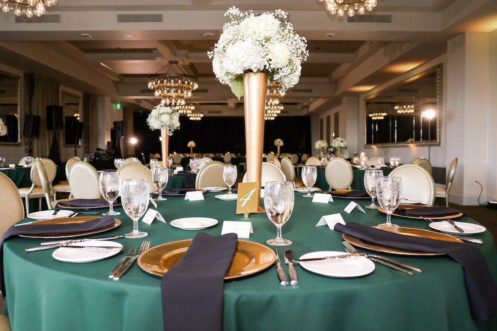 Old Hollywood Inspired Wedding Reception Decor, Round Tables with Emerald Green Tablecloths, Gold Chargers with Black Linens, Tall Gold Vase with Classic White Floral Arrangement Centerpiece and Gold Table Number Sign | Tampa Bay Wedding Photographer Lifelong Photography Studio | Downtown St. Pete Wedding Venue The Birchwood