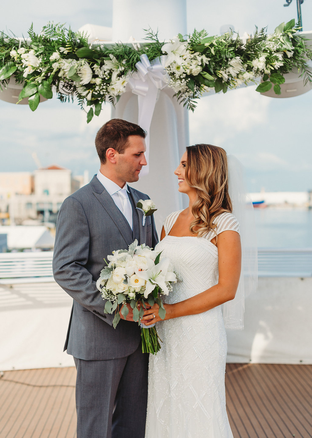 Florida Bride and Groom Wedding Portrait on Deck of Yacht, Bride in Scoop Neck Cap Sleeve Wedding Dress with Greenery, and White, Ivory Floral Bouquet, Groom in Grey Suit | Tampa Waterfront Wedding Venue Yacht Starship IV